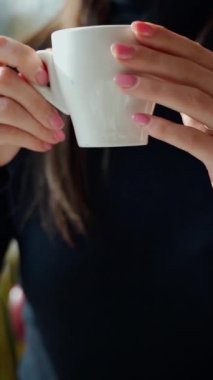 Kafede kahve içen güzel bir kız. Genç bir kadın pencerenin yanındaki masada oturuyor ve restoranda sıcak içkinin tadını çıkarıyor. Ağır çekim. Dikey video