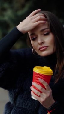 Parktaki şık bir kız. Güneşli bir günde, arka planda plastik bir fincan kahveyle kürklü güzel bir kadının portresi. Ağır çekim. Dikey video