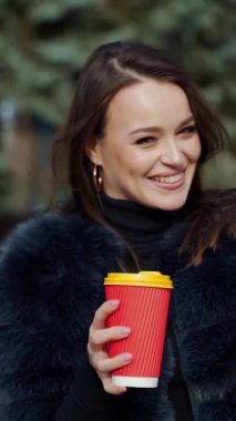 Kışın güneşli bir günde mutlu bir kızın portresi. Kürk mantolu güzel bir kadın parkta bir fincan sıcak içecek tutuyor ve kameraya gülümsüyor. Ağır çekim. Dikey video