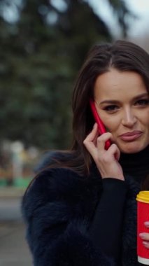 Telefonu olan güzel bir kadın ve bir fincan sıcak kahve ya da çay. Lüks kürklü güzel bir kızın portresi cep telefonuyla konuşuyor ve parkta kahve içiyor. Dikey video