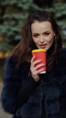 Açık havada bir modelin portresi. Plastik cam tutan kürklü zarif kadın parktaki kameraya bakıyor. Kahve içen alımlı kadının yüzü. Dikey video