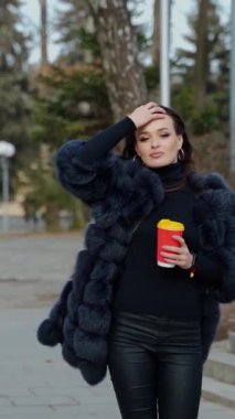 Şehir parkında kürklü güzel bir kız. Elinde bir fincan kahveyle dışarıdaki kameraya bakan muhteşem bir kadın manzarası. Ağır çekim. Dikey video