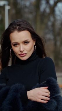 Güzel bir modelin portresi. Dışarıdaki kameraya poz veren siyah kürklü çekici kız. Parktaki çekici esmer kadın. Kamera hareket ediyor. Dikey video