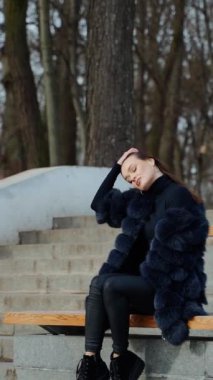 Dışarıda, yapraksız ağaçların arkasında oturan güzel bir kadın. Parkta dinlenen siyah kürklü seksi bir kadının yan görüntüsü. Ağır çekim. Dikey video