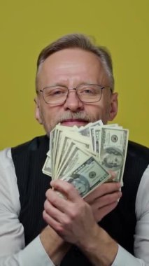 Dolar banknotları sallayan mutlu kıdemli işadamı. Elinde büyük miktarda para tutan ve kamerada gösteren zengin gri saçlı adam. Dikey video