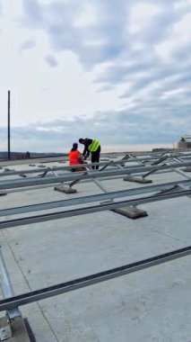 Düz bir çatıda gelecekteki güneş enerjisi çiftliği. Koruyucu üniformalı iki işçi fotovoltaik paneller yerleştirmek için metal taban kuruyor. Dikey video