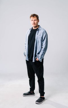 Stüdyo ortamındaki bir adamın sıradan portresi. Stüdyoda rahat bir şekilde duran, elleri ceplerinde sıradan bir kıyafet giyen, rahat bir tavır sergileyen bir adam..