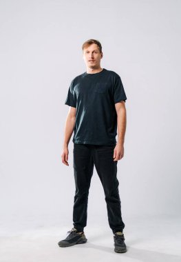 Stüdyo ortamında gelişigüzel duran genç bir adam. Genç bir adam minimalist bir stüdyoda rahat bir şekilde duruyor. Günlük siyah tişört ve siyah pantolon giyiyor.
