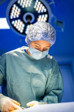 Steril ortamda çalışan bir cerrah. Ameliyat önlüğü ve maskeli bir cerrah titizlikle parlak ameliyat ışıkları altında bir hasta üzerinde çalışır.