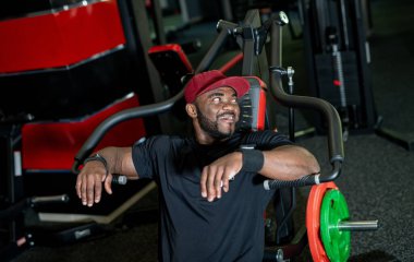 Spor salonundaki antrenmandan sonra rahatlayan gülümseyen adam. Kırmızı şapkalı formda bir adam, modern bir spor salonunda yoğun bir antrenmandan sonra bir anlık dinlenmenin keyfini çıkarır..