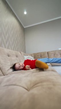 Neşeli, enerjik çocuk kanepede uzanıp bacaklarını sallıyor. Çocuk doğruldu ve yere düştü. Dikey video.