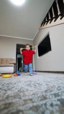 Kırmızı tişörtlü ve mavi pantolonlu beyaz erkek bebek odaya giriyor. Çocuk kameraya yakın bir oyuncak fırlatıyor, almaya geliyor ve kaçıyor. Düşük açı. Dikey video.