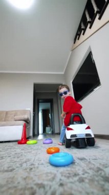 Güneş gözlüğü takan küçük bir çocuk oturma odasının yanında oyuncak bir arabaya biniyor. Evde oynayan çocuğun düşük açılı görüntüsü. Dikey video.