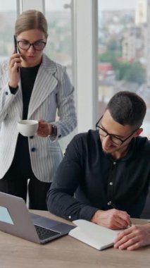 Koyu renk saçlı adam not defterine yazıyor ve dizüstü bilgisayarına bakıyor. Kadın meslektaşım telefonda konuşurken yanında duruyor ve kahve içiyor. Dikey video
