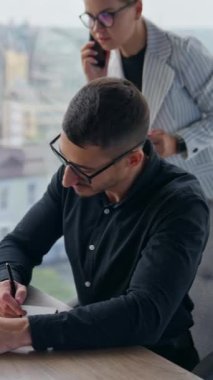 Çalışkan erkek çalışan not defterine not alıyor. Elinde bir fincan kahveyle pencerede duran kadın telefonda konuşuyor. Dikey video
