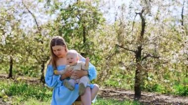 Mavi elbiseli uzun saçlı kadın dışarıda bebeğini kucağında tutuyor. Anne bahçede oğluna bir çiçek gösterir..