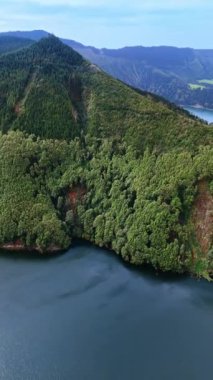 Yeşilliklerle kaplanmış bir dağın görüntüsü. Göl üzerindeki insansız hava aracı uçuşu dağlarda gerçekleşiyor. Azorean Adaları. Dikey video.