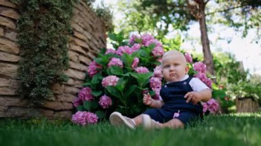 Mavi tulum giyen küçük sevimli çocuk çimlerin üzerinde oturuyor. Doğada oturan sarışın bebeğe alçaktan bir bakış açısı.
