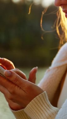 Beyaz ceketli, açık havada akıllı telefonunu kullanan beyaz saçlı bir kadın. Bulanık arkaplanda güneş ışınları.