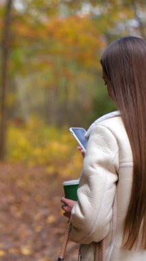 Uzun saçlı bir kadın elinde bir kağıt bardakla telefonuna bakarak yürür ve kameraya arkasını döner. Gülümseyen bayan arkasını döner ve birkaç adım geri gider. Arka planda sonbahar parkı.