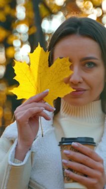 Elinde kağıt bardakla, sarı bir akçaağaç yaprağının arkasında yüzünün yarısını kaplayan güzel bir kadın. Kadın yaprağa bakar ve onu çevirir ve güneşe doğru yükseltir. Kapatın. Bulanık sonbahar arkaplanı.