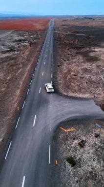 İki şeritli otoban, beyaz bir araba geçiyor. Asfalt yarış pisti İzlanda 'nın sürrealist düz arazisinden geçiyor. İnsansız hava aracı görüntüleri. Dikey video.