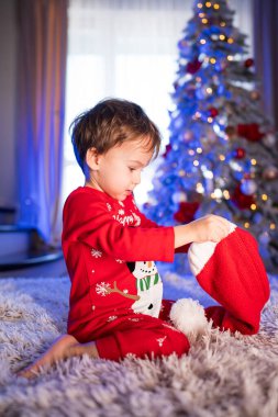 Ağacın yanında Noel çorabı açan bir çocuk. Şenlikli pijamalı bir çocuk, yumuşak ışıklarla parıldayan güzel dekore edilmiş bir ağacın yanında kırmızı bir Noel çorabını inceliyor..
