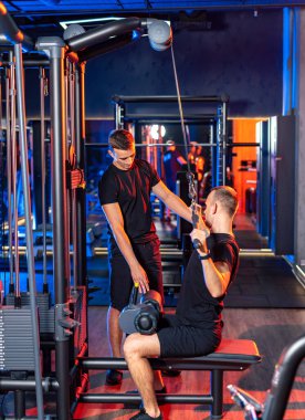 Spor aletleriyle çalışan iki adam. Bir fitness eğitmeni, kişisel antrenman sırasında modern bir spor salonunda kablo makinesi egzersizleri yapan bir müşteriye yardımcı olur..