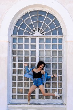 Oyuncu kadın gündüz vitrininde poz veriyor. Genç bir kadın, güneşli bir günde büyük bir pencereden sarkan şık bir kıyafetle poz verirken sevinç ifade eder..