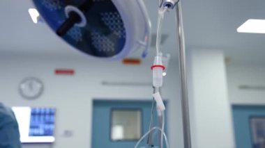 Damardan damlayan odak noktası ilaç şişesine bağlı. Sıhhiye grubu arka plandaki yuvarlak lambanın altında bulanık bir şekilde çalışıyor..