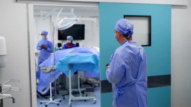 Mavi üniformalı erkek cerrah ameliyathanenin açık kapısında duruyor. Ameliyat masasında ameliyat için hazır bekleyen iki doktor var..