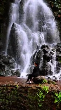 Esmer kadın taşın üzerinde oturuyor ve güzel şelaleye bakıyor. Kara kayaların oradan su fışkırıyor. Azorean Adaları 'nın doğası. Dikey video.