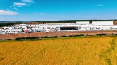 Büyük sanayi fabrikasının önündeki sarı tarla. Drone fabrikaya yaklaşıyor. Önünde büyük bir otopark var..