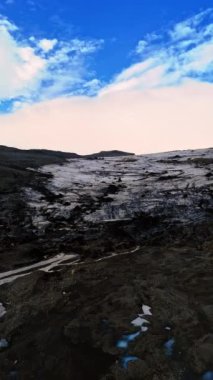Nehirleri akan siyah dağlık bir arazi. Antik eriyen buzullar dağın tepesinde. Arka planda beyaz bulutlu mavi gökyüzü. Dikey video.
