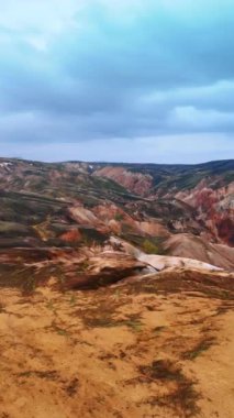 Gökyüzünden İzlanda 'nın sürrealist rengarenk dağlık manzarası. Bulutlu gökyüzünün arka planında çorak kayalar ve vadiler. Dikey video.