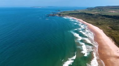 İrlanda sahillerindeki kumlu sahilde beyaz dalgalar akıyor. Güneşli manzaralı güzel bir su manzarası ve tepeden yeşil bir kıyı..