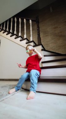 Kırmızı tişört giyen bebek evdeki merdivenlerde oturuyor. Çocuk bir şişe alıyor ve ondan içiyor. Düşük açı. Dikey video.