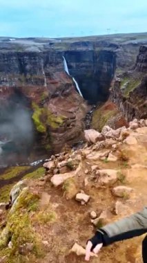Tehlikeli kayanın kenarında duran bir çifte yaklaşıyoruz. İnsansız hava aracı dağlardaki oluğa uçarak kayaların arasından akan muhteşem şelalenin ve nehrin manzarasını gözler önüne seriyor. İzlanda. Dikey video.
