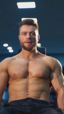 Spor salonunda yakışıklı bir sporcunun portresi. Genç sakallı, gömleksiz, sandalyede oturan ve kol kaslarını ağır dambıllarla pompalayan adam. Dikey video