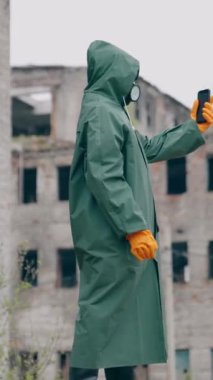 Koruyucu giysili bir insan ve telefonlu bir gaz maskesi. Güvenlik üniformalı, eldivenli ve solunum cihazlı tanınmayan biri radyasyon sırasında zarar görmüş yerde selfie çekiyor. Dikey video