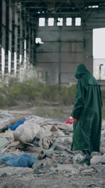 Terk edilmiş binada koruyucu giysili ve solunum cihazlı bir adam var. Mahvolmuş yerdeki çöp yığınının yanında güvenlik kıyafetli bir ekolojist. Radyasyon bölgesi. Dikey video