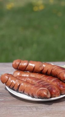 Dışarıdaki masada sebzeli ızgara sosis hazır. Şef bir tabağa kızarmış sosis koyar. Piknik için lezzetli bir yemek. Dikey video