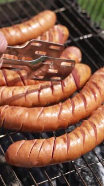 Kömürde ızgarada kızarmış sosis. Aşçı, yaz mevsiminde bahçede kızarmış sosis ve tong ile döner. Piknik için lezzetli yemekler. Dikey video