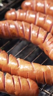 Izgarada lezzetli sosisler. Aşçı piknik için neredeyse hazır sosisleri çeviriyor. Bahçedeki kömürlerin üstünde yemek kızartıyorlar. Yakın plan. Dikey video