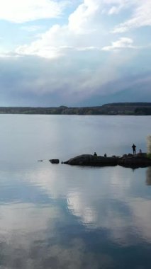 İlkbaharda doğa manzarasında güzel bir nehir. İlkbaharda insanlar taşların üzerinde suyun yanında dinlenirler. Nehrin kıyısında çiçek açan ağaçlar. Dikey video