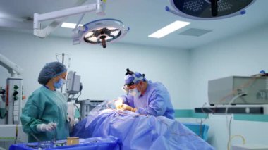 Erkek cerrah cerrahi aletlerle hastanın üzerine eğildi. Kadın ameliyat hemşiresi aletleri doktora verir..