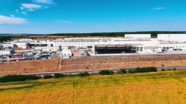 İnsansız hava aracı görüntüleri, kırsal kesimdeki modern bir girişimin geniş bir bölgesinden uzaklaşıyor. Otobüsler, kamyonlar ve arabalar fabrikanın önündeki otoparkta duruyor..