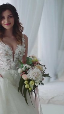 Yatak odasında genç ve güzel bir gelin. Beyaz gelinlik içinde güzel genç gelin. Dikey video.