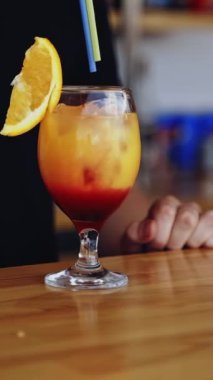 Bardakta lezzetli bir kokteyl. Barmen müşteri için içecek pişirir. Bar masasında taze alkol içeceği hazır. Bardakta portakal dilimi, buzlu renkli sıvı. Dikey video