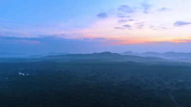 Gün batımında puslu manzaranın üzerindeki görüntüler. Arka planda dağların çoklu bitki örtüsü ve siluetleri olan bir düzlük. Hava görünümü.